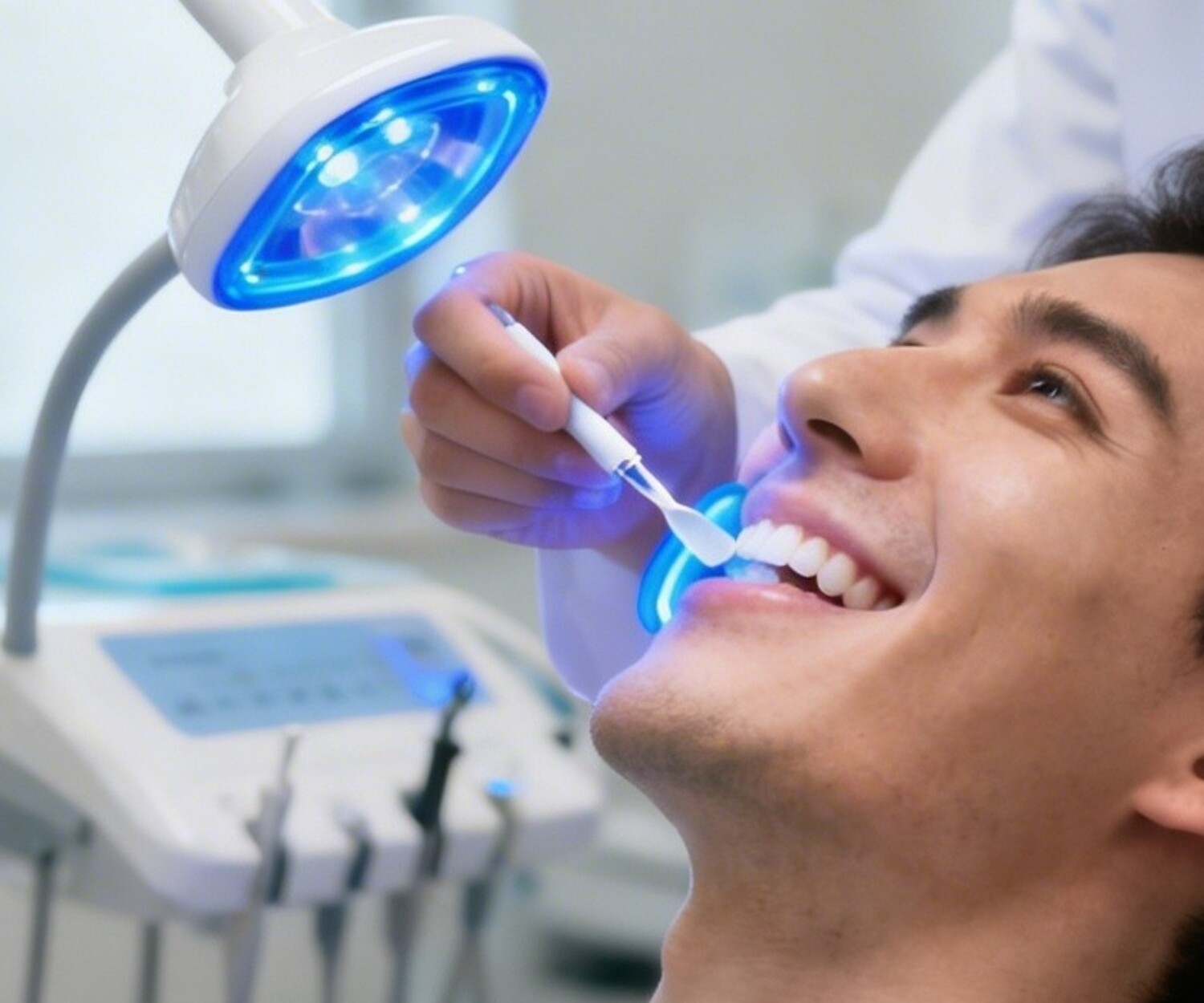Bright, modern dental clinic scene showing a teeth whitening procedure in progress. Patient sitting in a dental chair with mouth open while a dentist applies whitening gel and uses an LED whitening light on the teeth.