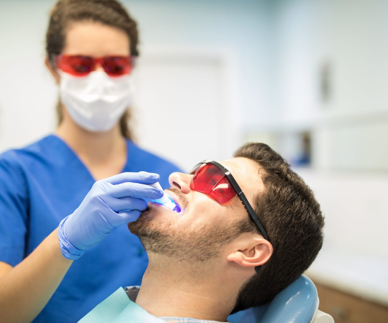 Dentist performing laser teeth whitening treatment on a male patient wearing protective glasses in a dental clinic.